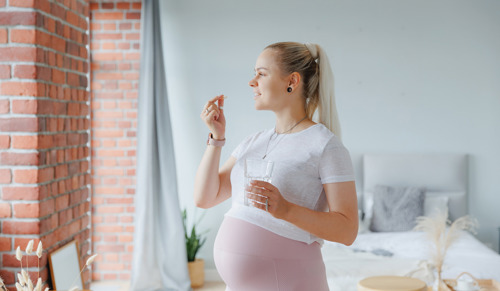 Sretna mlada trudnica s bijelom okruglom pilulom i čašom vode u ruci, dnevni vitamini za kosu, kožu i zdravlje bebe